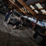 In einer Garagenhalle steht eine Frau präsentabel vor der Motorhaube eines Geländewagens, während der Mann links an das Auto lehnt