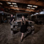 In einer Garagenhalle steht eine Frau präsentabel vor der Motorhaube eines Geländewagens, während der Mann links an das Auto lehnt