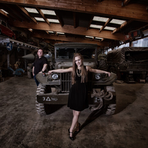 In einer Garagenhalle steht eine Frau präsentabel vor der Motorhaube eines Geländewagens, während der Mann links an das Auto lehnt